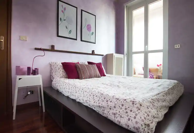 Camera da letto luminosa e di stile con un letto su piattaforma basso, biancheria floreale e accenti viola coordinati. Ampia finestra/porta illumina la stanza e si intravede un piccolo angolo salotto.