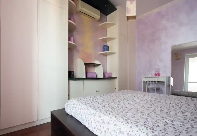 Camera da letto luminosa dai toni pastello con un grande letto in primo piano, armadi a muro e mensole, e un piccolo trucco vicino alla finestra.