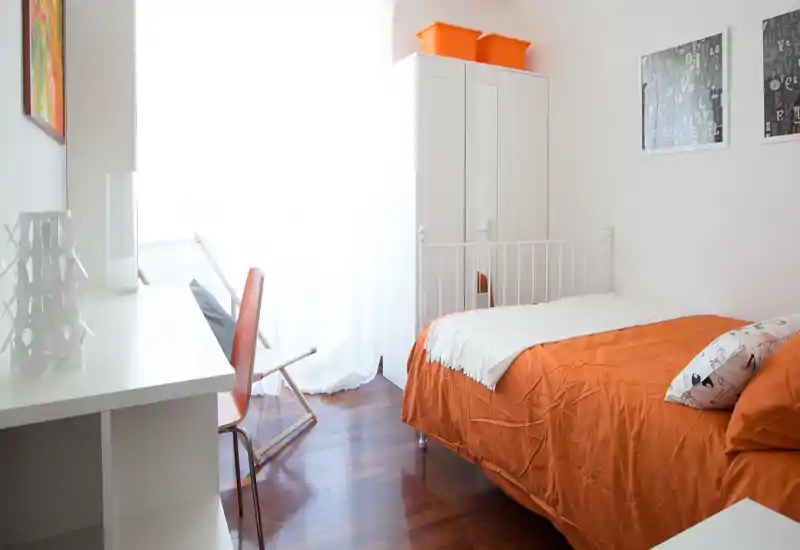 Camera singola luminosa e accogliente con biancheria arancione, armadio bianco con specchio, scrivania e luce naturale da una grande finestra. Stile pulito e moderno, ideale per un annuncio.