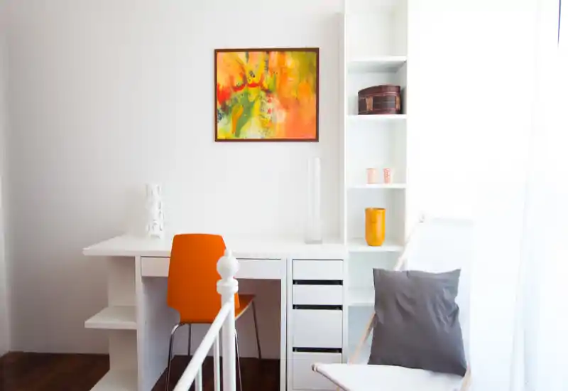 Angolo camera luminoso e minimale con scrivania bianca, sedia arancione e un accenno alla pediera del letto in primo piano; scaffalatura con oggetti decorativi e un quadro colorato sopra la scrivania donano un aspetto moderno e arioso.