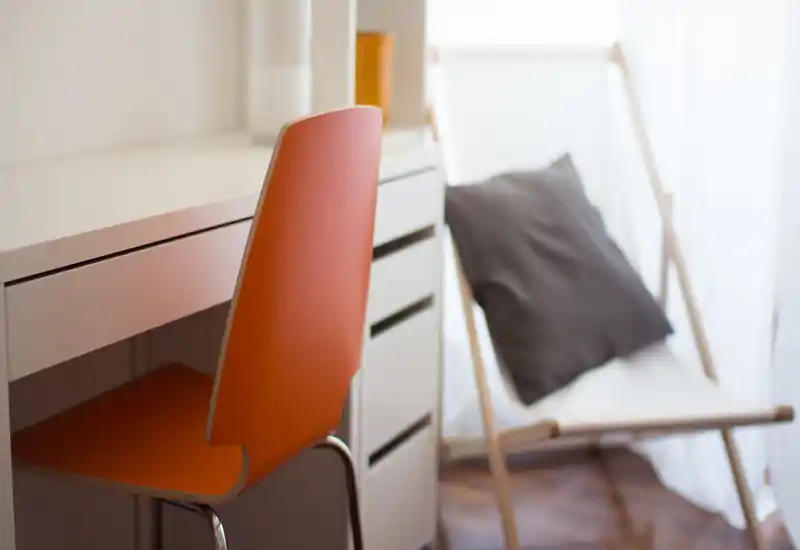 Angolo luminoso e moderno con una scrivania bianca e una sedia arancione in primo piano e una poltrona relax con cuscino vicino alla finestra.