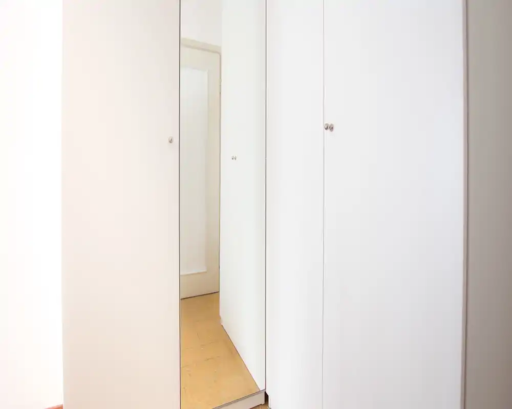 Primer plano de puertas de armario blancas con un estrecho panel espejo que refleja una puerta de pasillo y el suelo; información contextual mínima sobre la habitación circundante.