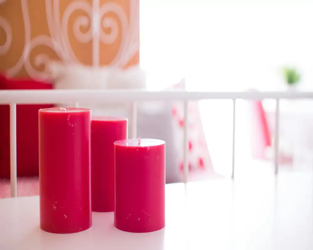 Detalle luminoso y suave de dormitorio con velas decorativas rojas en primer plano y cabecero metálico blanco y cojines en un fondo desenfocado: composición acogedora perfecta para fotos de alquiler.