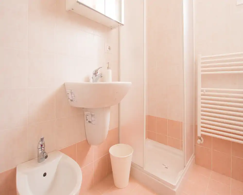 Baño compacto y limpio con ducha en esquina, lavabo pared y bidé, azulejos color melocotón claro y toallero calefactado.