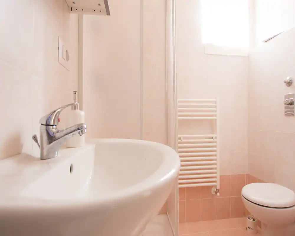 Baño pequeño y luminoso con lavabo blanco, grifo cromado, inodoro y toallero calefactado; azulejos claros y luz natural que aportan sensación de limpieza y amplitud.