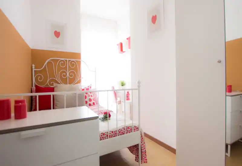 Dormitorio pequeño y luminoso con cama de metal blanco, ropa de cama estampada y pared en tono ocre; la luz natural suave crea un ambiente acogedor.