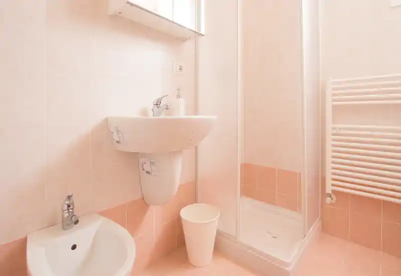 Salle de bain compacte et propre avec cabine de douche d'angle, lavabo mural et bidet, carrelage pêche clair et sèche-serviettes.