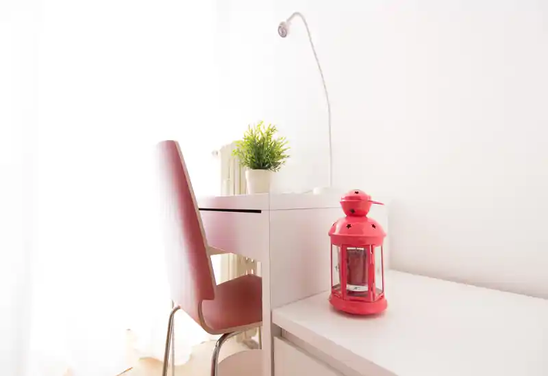 Rincón luminoso y minimalista de la sala con escritorio blanco, silla roja y linterna decorativa; la luz natural y una planta pequeña hacen el espacio acogedor.