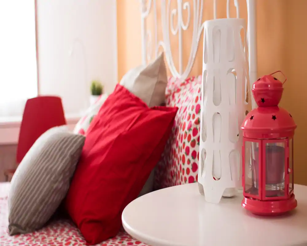 Bright, cozy bedroom corner with patterned bedding and multiple decorative pillows; a small bedside table with a modern white lamp and a pink lantern adds charming detail.