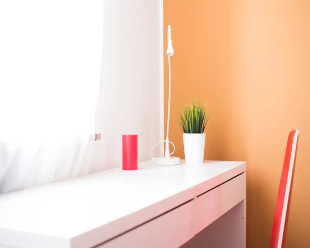 Bright, minimal desk corner with a small white lamp, potted plant and a colorful orange wall creating a cozy reading/work spot.