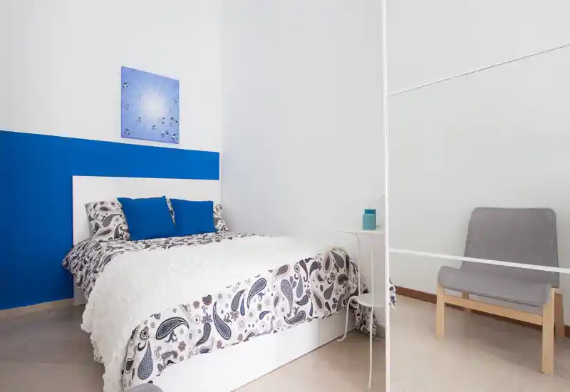 Dormitorio luminoso y ordenado con edredón estampado, pared de acento azul y cojines a juego. La cama está bien encuadrada y el espacio transmite sensación de frescura y confort.