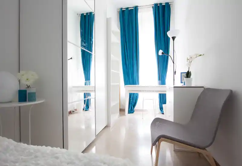 Dormitorio moderno y luminoso con muebles blancos, cama en primer plano, cortinas azules de suelo a techo y armario con puertas espejo que amplía el espacio.