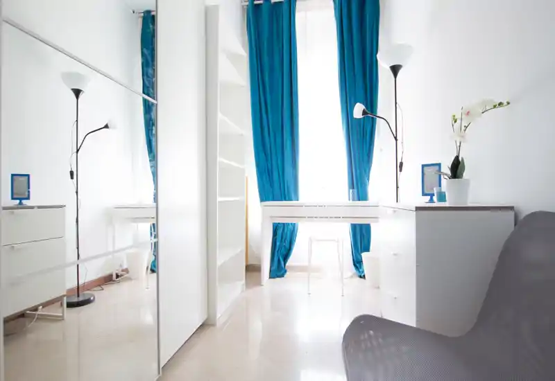 Zona de estar moderna y luminosa con mobiliario blanco, cortinas turquesa y un pequeño escritorio junto a una gran ventana que aporta abundante luz natural.