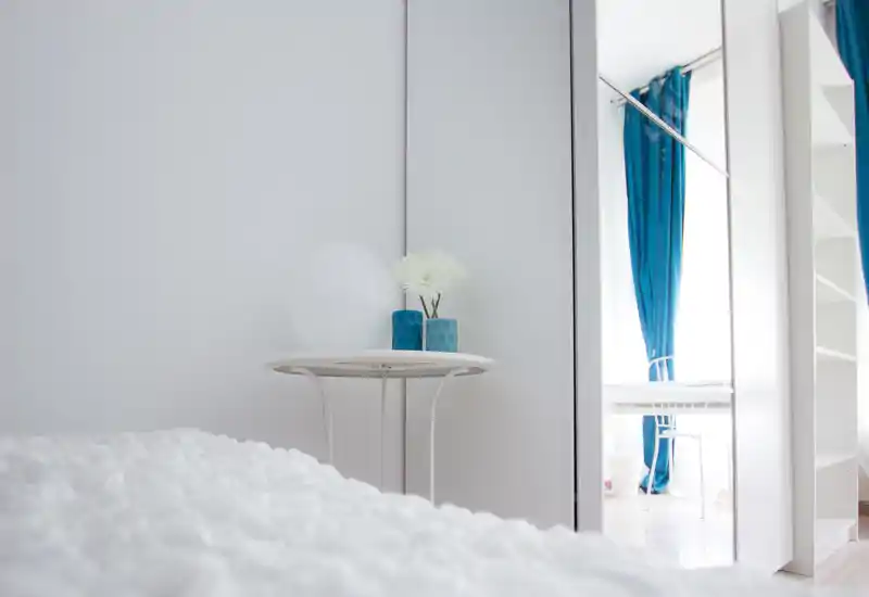 Dormitorio minimalista y luminoso con edredón blanco texturado, mesita redonda con jarrones decorativos; armario con espejo y cortinas azules que añaden profundidad y contraste.