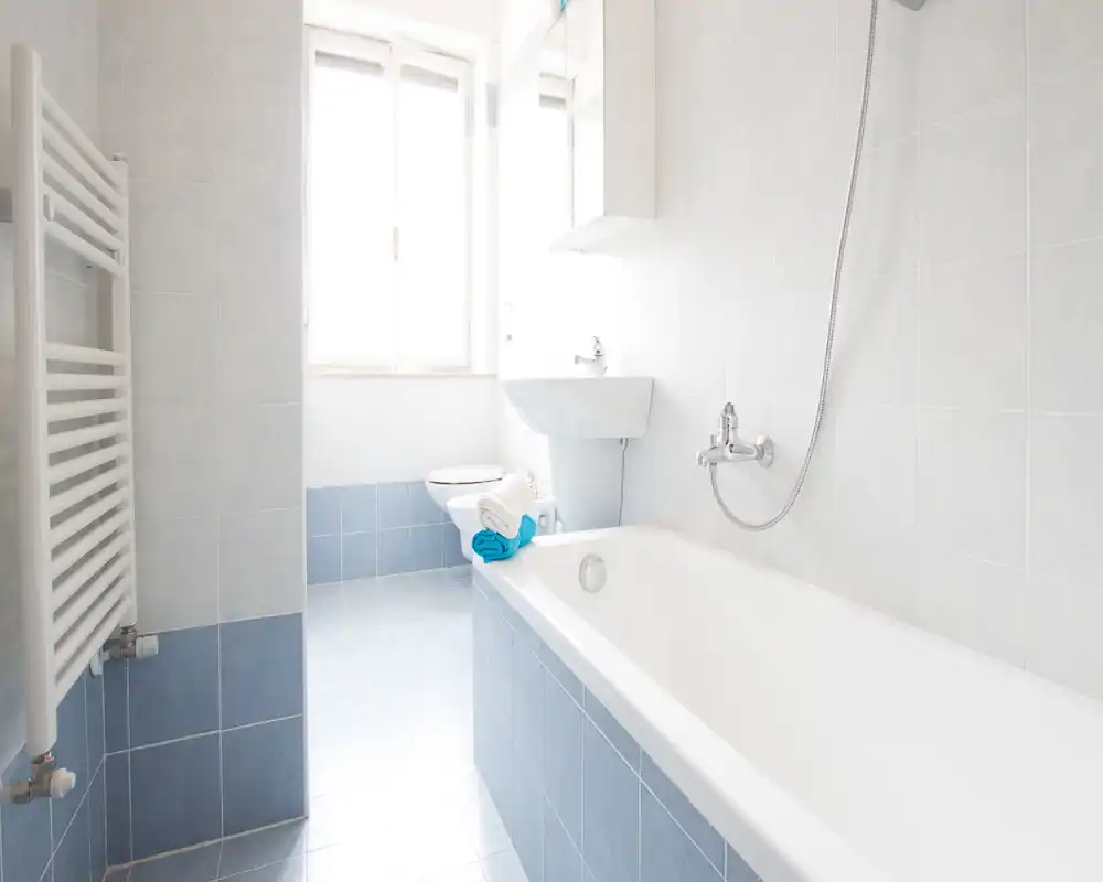 Baño luminoso y limpio con bañera, grifo mural y ducha de mano, lavabo exento y WC. Azulejos azul claro y luz natural desde la ventana que crean una sensación de amplitud.