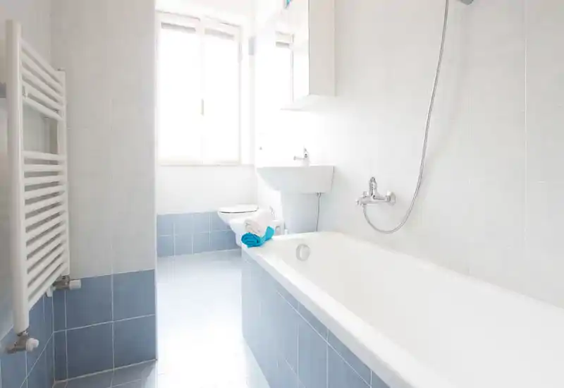 Salle de bains lumineuse et propre avec baignoire, robinet mural et douchette, lavabo sur pied et WC. Carrelage bleu clair et lumière naturelle provenant de la fenêtre pour une ambiance aérée.