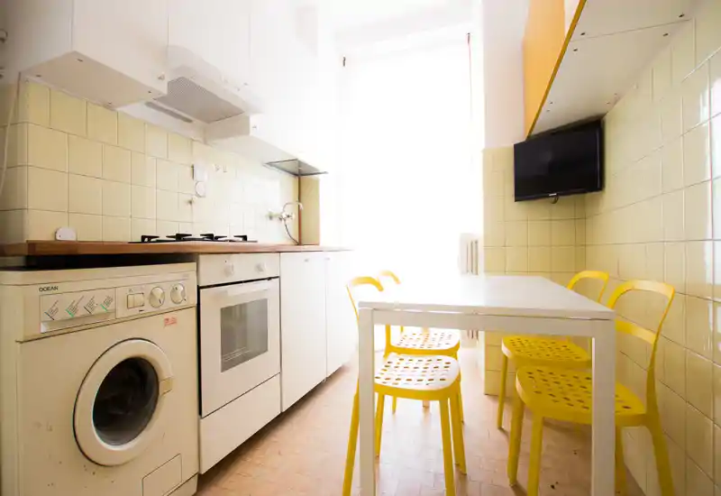 Cuisine compacte et lumineuse avec placards blancs, plaque de cuisson à gaz et four encastré, lave-linge et une table avec quatre chaises jaunes; grande fenêtre apportant une forte lumière naturelle.