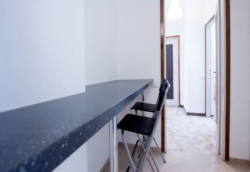 Bar kitchenette étroit avec deux tabourets hauts et un long plan de travail près du couloir. Espace propre et minimal, adapté aux repas rapides ou à un studio compact.