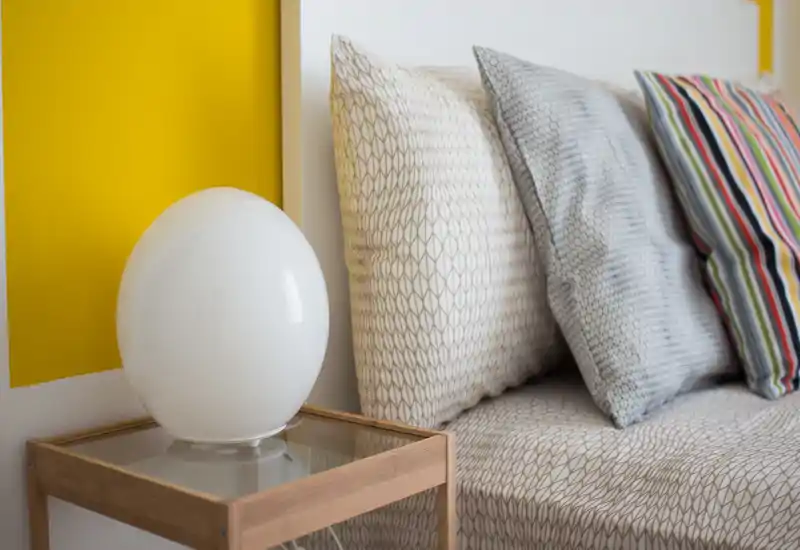 Plano cercano de un rincón de dormitorio bien decorado con almohadas, cabecero y mesita de noche con lámpara moderna; decoración contemporánea y luminosa.