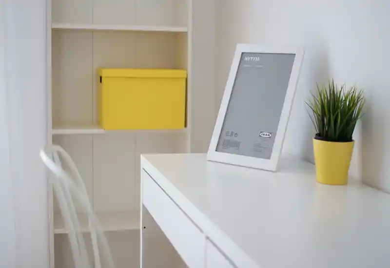 Bright, minimal living area corner with a white desk, decorative frame and a small potted plant in a yellow pot; clean Scandinavian styling.