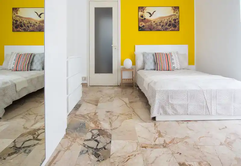 Dormitorio moderno y luminoso con cama individual frente a una pared amarilla vibrante, cojines decorativos, mesita y cuadro. Suelo de mármol y gran espejo aportan profundidad y luz.