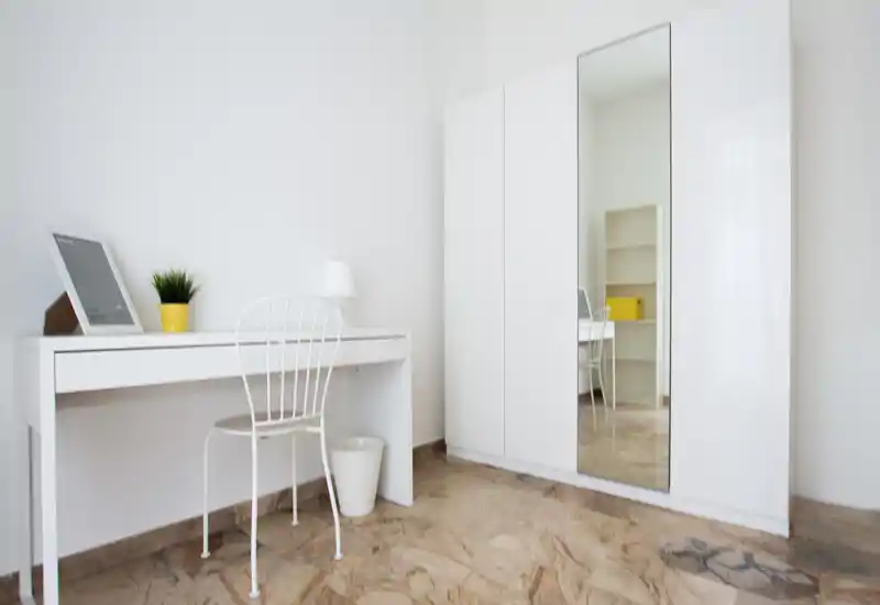 Rincón de dormitorio minimalista con armario blanco (una puerta con espejo), escritorio compacto y silla blanca, suelo efecto mármol; luminoso y despejado, ideal para una persona.