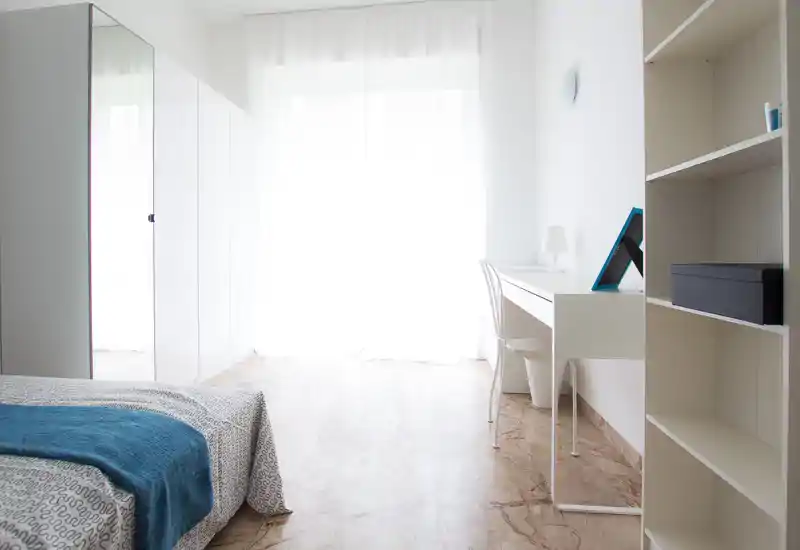 Dormitorio luminoso y minimalista con cama parcialmente visible, escritorio blanco junto a la ventana y armario brillante que refleja la luz — ideal para anuncios que destacan la luz natural y el diseño limpio.