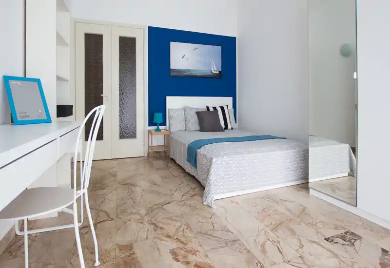 Dormitorio moderno y luminoso con cama doble frente a una pared azul intensa, cojines decorativos y una pequeña lámpara de noche. Escritorio y silla a la izquierda y armario con espejo que amplía la sensación de espacio.