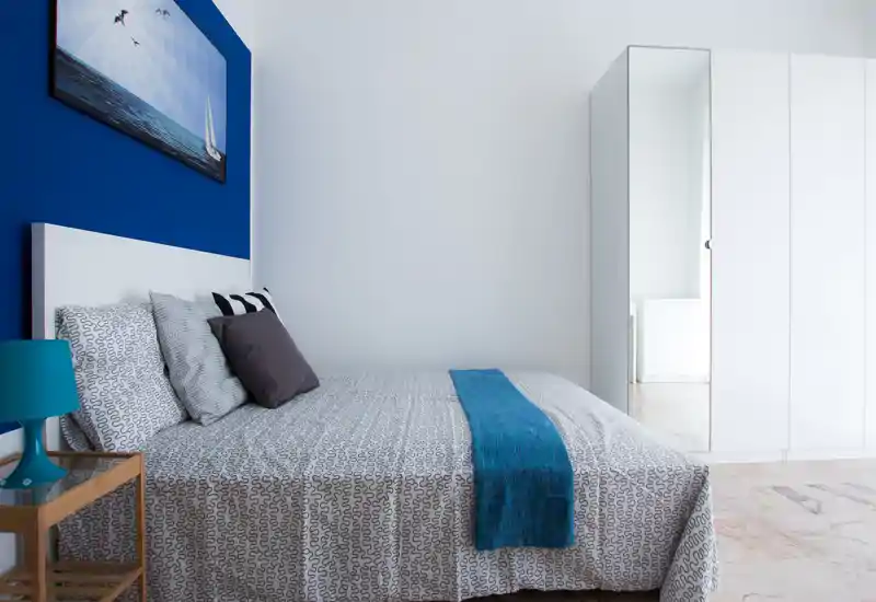 Dormitorio moderno y luminoso con cama doble frente a una pared azul intensa, ropa de cama estampada y una franja azul. Un armario blanco con puerta espejo y una mesita con lámpara completan el ambiente minimalista y acogedor.