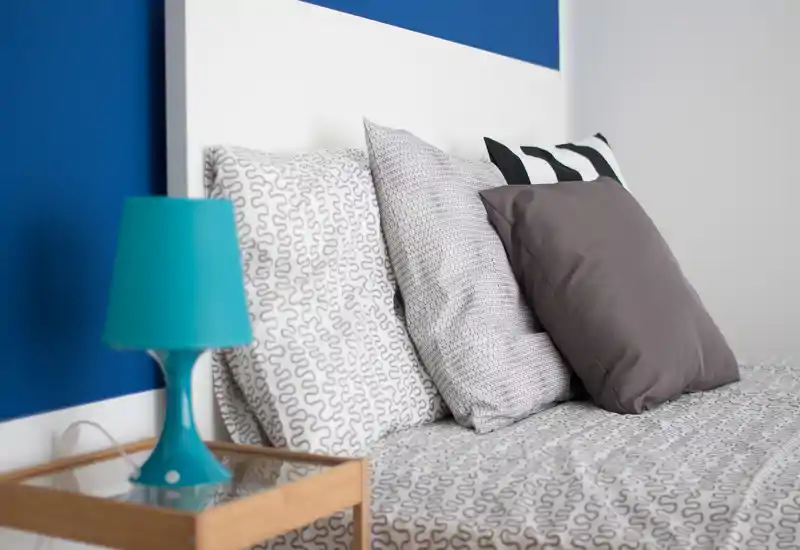 Primer plano de dormitorio elegante con cama bien hecha, sábanas estampadas, cojines superpuestos y una lámpara turquesa en la mesita frente a una pared azul.