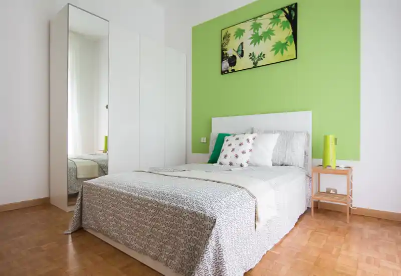 Dormitorio moderno y luminoso con cama doble, cojines decorativos y armario con puerta espejo. La pared verde y la obra de arte aportan color fresco, y el suelo de parquet hace la estancia cálida y acogedora.