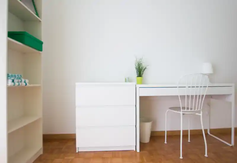Rincón de dormitorio luminoso y minimalista con mobiliario blanco, pequeño escritorio, silla y estantería — ordenado y aireado para un anuncio moderno.