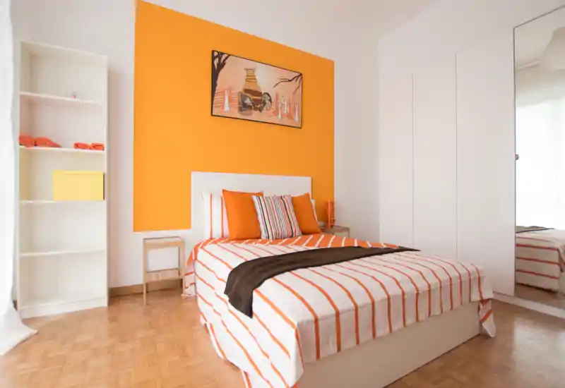 Bright, modern bedroom with orange-accented wall, striped bedding and wardrobe with mirrored door; comfortable double bed and simple shelving create a fresh, inviting space.