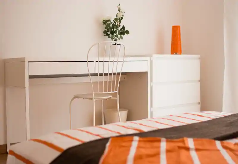 Bright, tidy bedroom corner with a visible bed foreground, white desk and chair, warm orange accents and minimal decorative touches.