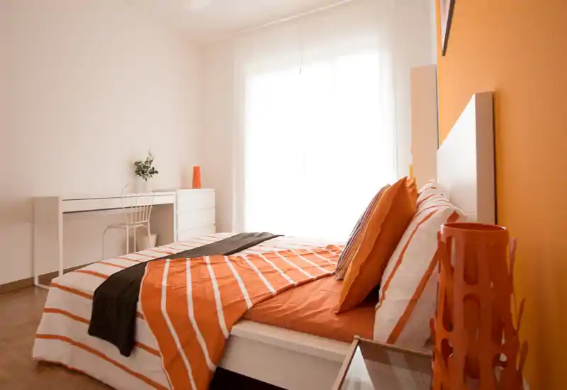 Bright, modern bedroom with orange-accented bed linens, pillows and bedside decor. Large window provides abundant natural light and a minimal desk area at the far wall.