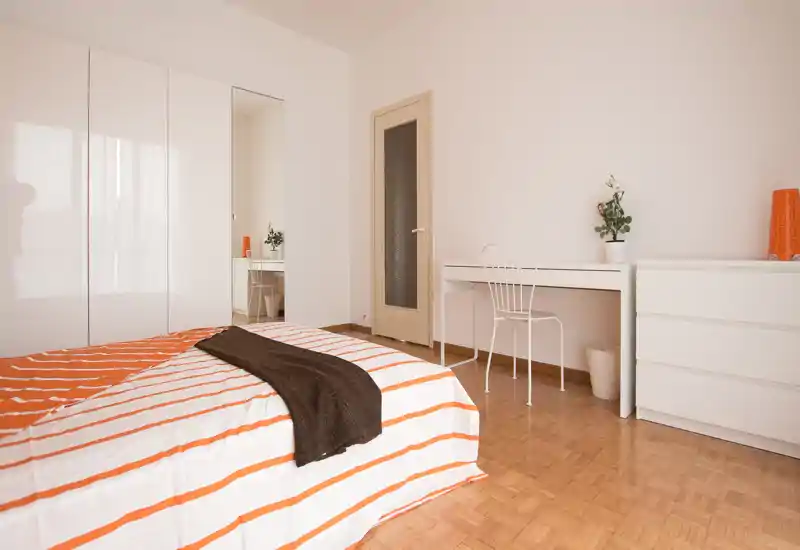Bright, minimalist bedroom with a striped bed, white furniture and warm parquet flooring — ideal for a clean, modern rental listing.