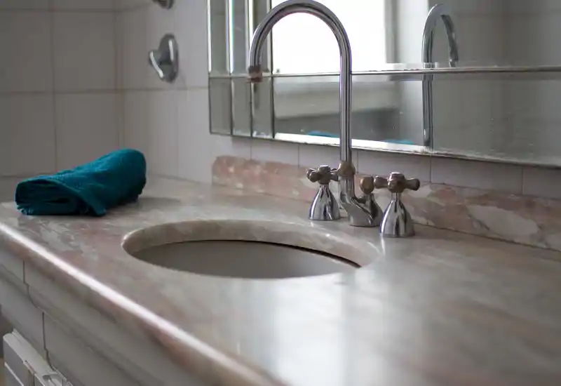 Close-up of a bathroom sink with marble countertop, chrome faucet and a folded teal towel on the left. The image highlights fixtures and countertop detail.