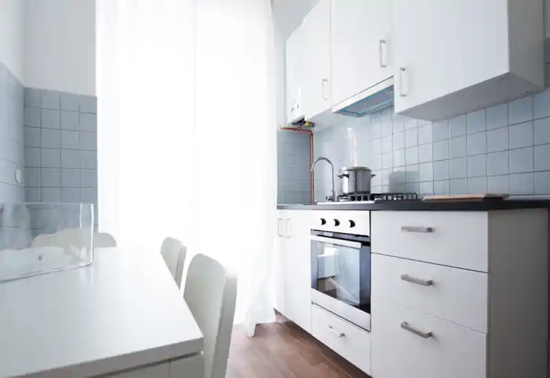 Cocina compacta y luminosa con muebles blancos, horno integrado y vitrocerámica de gas, con pequeña zona de comedor — moderna, minimalista y bien iluminada.