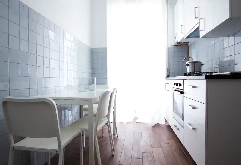 Cocina minimalista y luminosa con una mesa pequeña y cuatro sillas blancas, azulejos azul claro y suelo de madera—bien iluminada por una gran ventana.