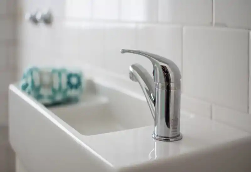 Close-up of a modern chrome faucet and white sink in a bright, clean bathroom with tiled backsplash and a folded towel in the blurred background.