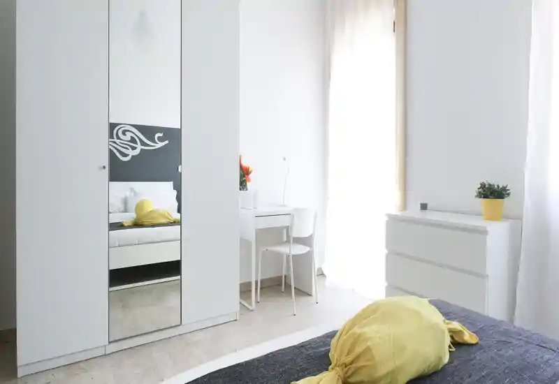 Dormitorio luminoso y minimalista con cama doble visible, armario blanco con espejo, pequeño escritorio junto a la ventana y luz natural suave.