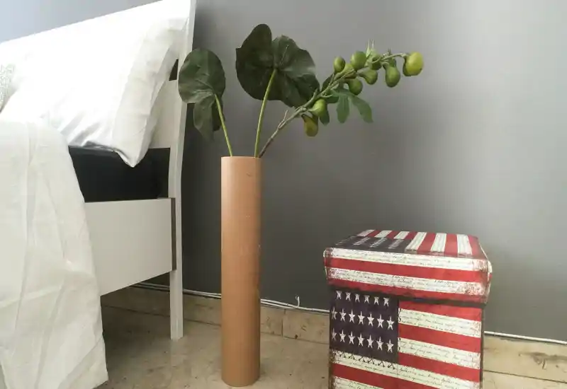 Rincón de dormitorio tranquilo con estructura de cama blanca, jarrón alto decorativo con follaje verde y un taburete rústico con estampado de la bandera estadounidense — luminoso, sencillo y adecuado para un anuncio.