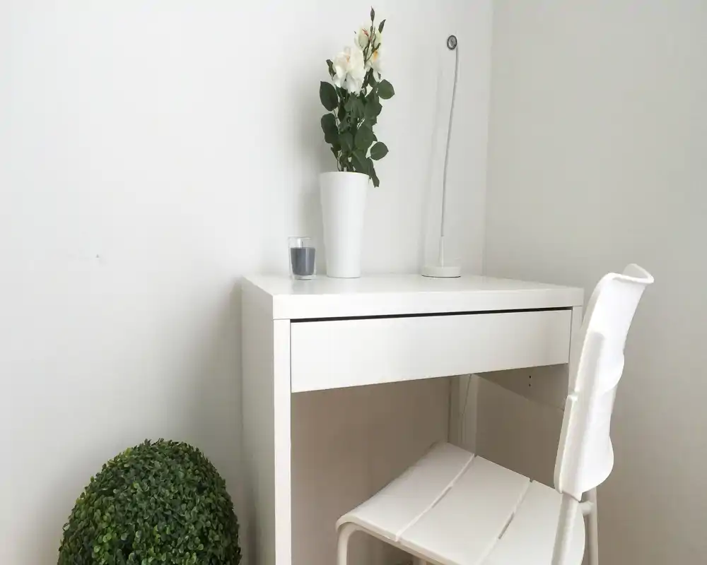 Rincón minimalista y luminoso con escritorio blanco y silla a juego, decorado con un jarrón de flores y una pequeña planta — ideal como espacio de trabajo compacto.