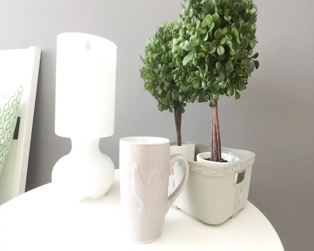Plano cercano de una mesita con lámpara blanca, plantas decorativas en maceta y una taza sobre una mesa clara, detalle minimalista y sereno del dormitorio.