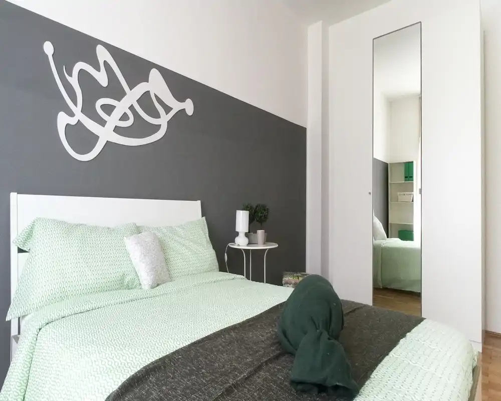 Dormitorio luminoso y bien preparado con cama en ropa de cama verde claro, un panel decorativo y armario con espejo que aporta profundidad.