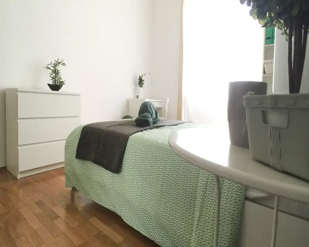 Dormitorio luminoso y minimalista con cama bien hecha en ropa de cama verde, suelo de madera y mobiliario blanco sencillo que transmite una sensación fresca y aireada.