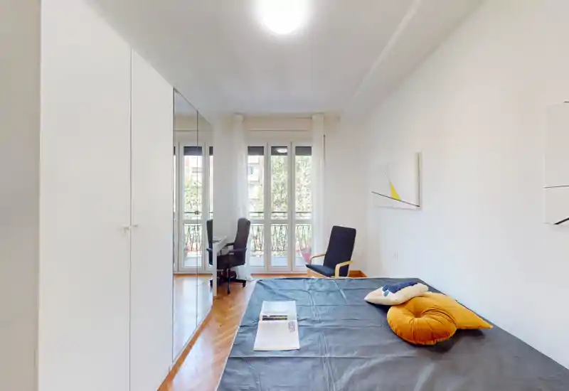 Dormitorio moderno y luminoso con una cama grande, cojines decorativos y un balcón que aporta luz natural. La habitación cuenta con un armario con espejo y un rincón con silla/escritorio, espacio funcional y ventilado.