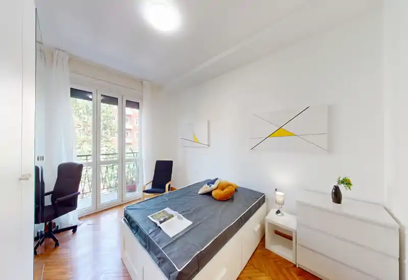 Dormitorio moderno y luminoso con cama centrada, suelo de parquet y amplias puertas al balcón que aportan mucha luz natural. Mobiliario blanco minimalista y una pequeña zona de trabajo, ideal para alquileres temporales o estudiantes.