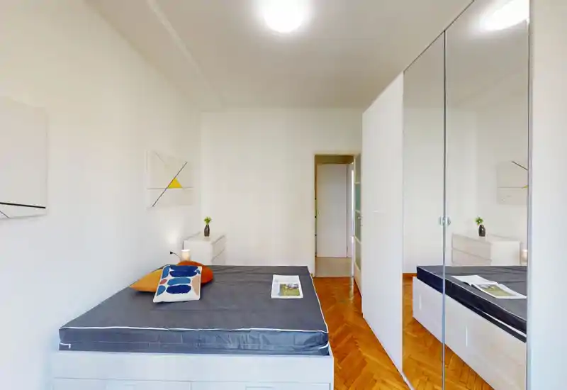 Dormitorio luminoso y minimalista con una cama grande, mesitas sencillas y un armario con espejos que aporta profundidad. Suelo de parquet y paredes blancas para un aspecto moderno y limpio.
