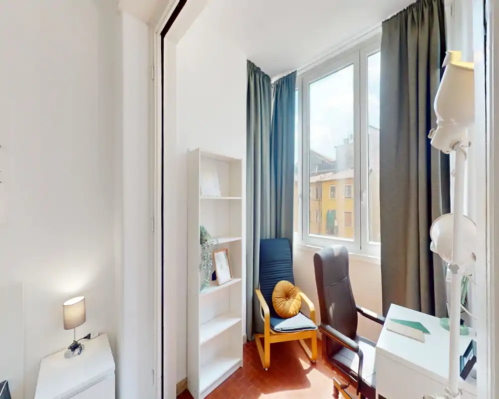 Zona giorno luminosa e compatta con due poltrone confortevoli vicino a una grande finestra, tende morbide e una piccola libreria — ideale per leggere o lavorare con molta luce naturale.